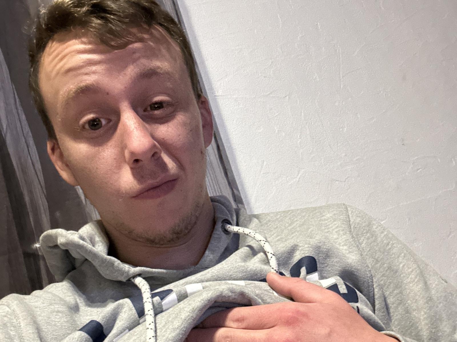 JojoGojo, a white dude with short brown hair, light stubble, and a goofy expression, wearing a grey hoodie, is holding his chest with a slightly open mouth.