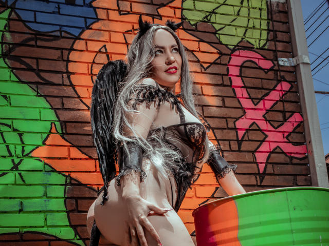 NatashaCoop, avec ses longs cheveux argentés, ses cornes de diable noires et ses ailes, portant de la lingerie en dentelle noire, montrant ses fesses, devant un mur coloré avec des graffitis, avec un seau vert et orange..