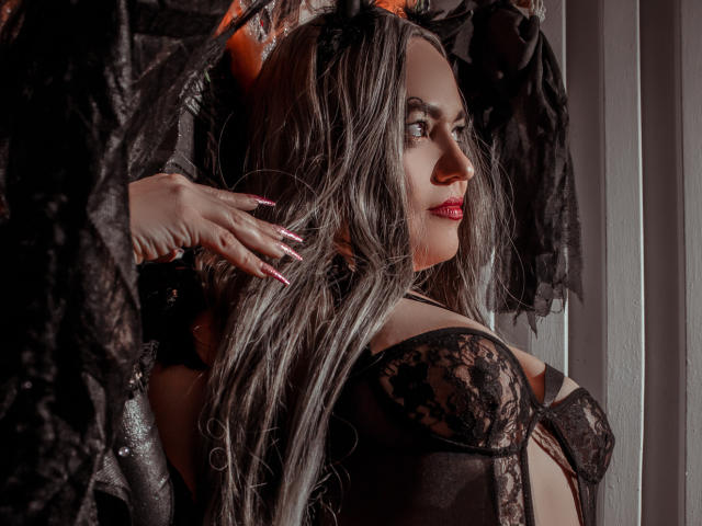 NatashaCoop, con cabello largo y gris, un sujetador de encaje negro y lápiz labial rojo, y con orejas de gato, tiene una mano en el pelo, mirando hacia un lado, mostrando sus pechos..