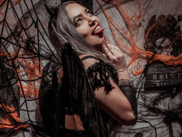 NatashaCoop, con cabello gris, orejas de gato negras y una expresión vivaz, con la lengua afuera, lamiendo un dedo, vestida con encaje negro, alas y cuero, mirando hacia atrás con una vibra juguetona, sexy y de bruja oscura..