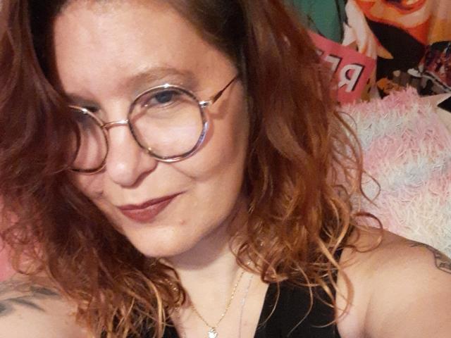 Choukette, con cabello rojo rizado, gafas redondas y lápiz labial oscuro, sonríe a la cámara, mostrando tatuajes en su brazo, y lleva una blusa negra. Su zona genital no es visible..