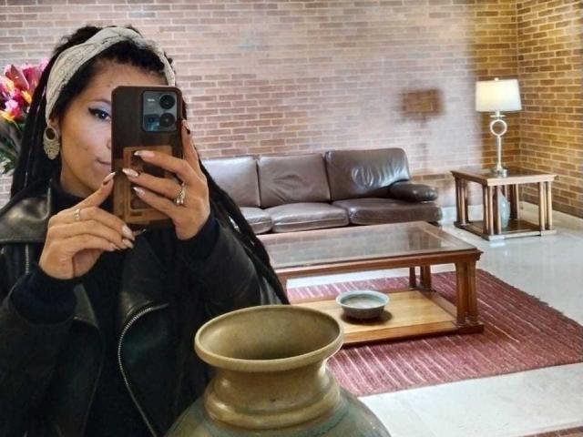 AmelieBlake with dark skin, wearing a gray headscarf and black leather jacket, taking a selfie with a phone, showing off her nails and ring, in a living room.