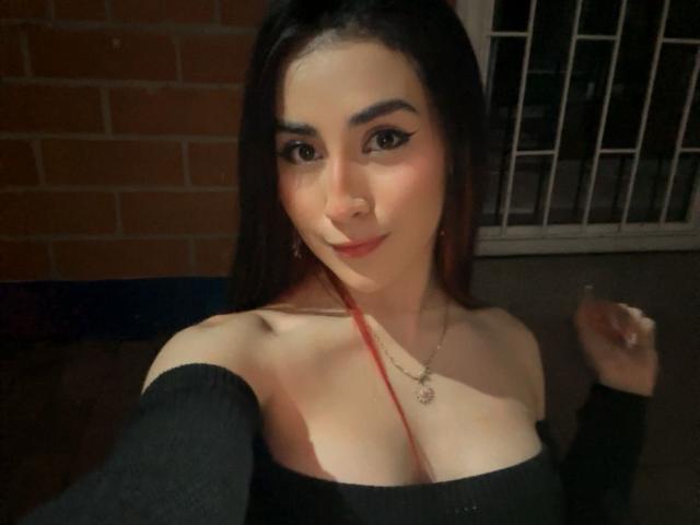AnyaJay has medium tits, dark hair, and is wearing a black off-shoulder top with a red straw, smiling at the camera, holding a cigarette.