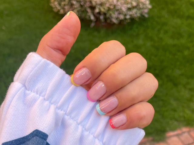 LunaMiranda's hand with glossy pink nail polish and colorful tips (yellow, blue, pink, red), wearing a white, puffy-sleeved shirt, pointing with her index finger.