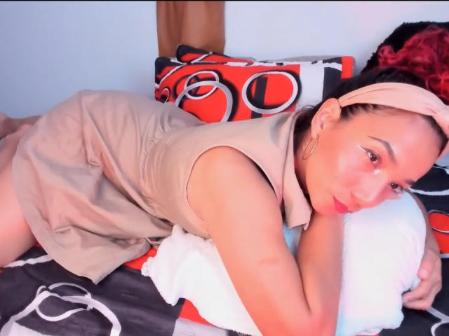 AmberDemons with tan dress, lying on bed, tan headband, red lipstick, small tits, brown skin, looking at camera, holding white pillow, red and black patterned pillows in background.