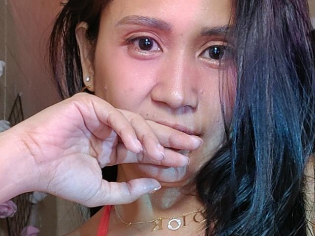 YaruShitai69 with wet black hair, pinkish skin, and small nose, holding her chin with her hand, wearing a gold necklace, and pearl earrings. Her face is slightly sweaty.