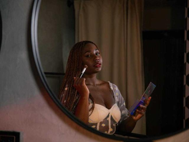 AkaneCox with dark skin, braided hair, holding a makeup brush to her cheek, wearing a low-cut white top, and holding a blue container, reflected in a round mirror.