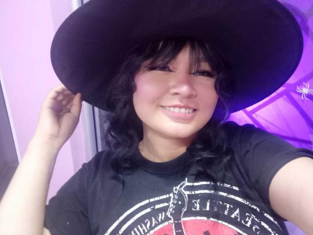 LillyMayer with wavy black hair, wearing a large black hat, smiling, touching her hat with her right hand, wearing a black T-shirt, and showing her teeth.