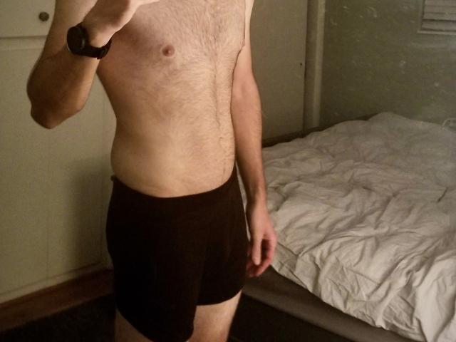 MikeBm, shirtless, wearing black boxer shorts, has a hairy chest, slim build, and a black wristwatch, standing in a dimly lit room with a bed on the right.