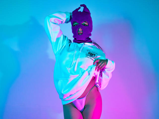 NaiaSanders wearing a purple mask, white hoodie, and pink thong, with one hand on her head and the other on her hip, under blue and pink lighting.