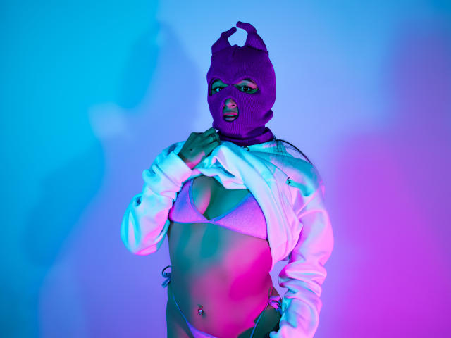 NaiaSanders with purple mask, white hoodie, lifting it to show pink bikini top, exposing her tits and belly button piercing, under blue and pink neon light.