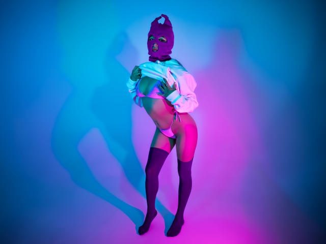 NaiaSanders in a purple balaclava, white shirt lifted to expose her tits, pink and white panties, and black thigh-highs, standing in colorful neon light, shadow on the wall.