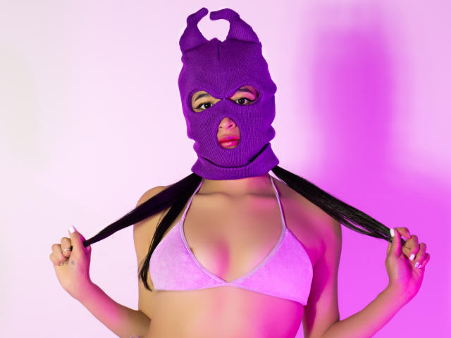 NaiaSanders wearing a purple mask with horns, holding black pigtails, in a white bikini top, pink and purple lighting. Boobs, mask covers nose and mouth.