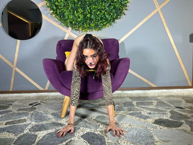RomantikHotCouple, with wavy brown hair and dark lipstick, crouches on a purple chair, hands on stone floor, leopard print top, black panties, yellow underwear peeking, looking at camera.