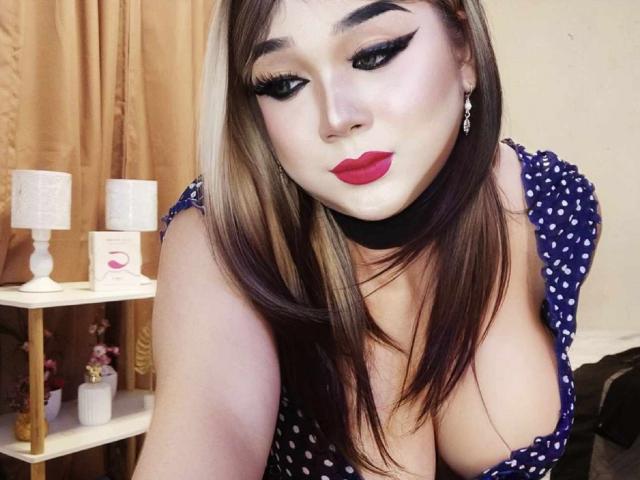 MissBigCockLucy with big tits, dark eyebrows, red lipstick, black choker, long brown hair, and black eyeliner, wearing a blue polka dot dress, in a bedroom selfie.