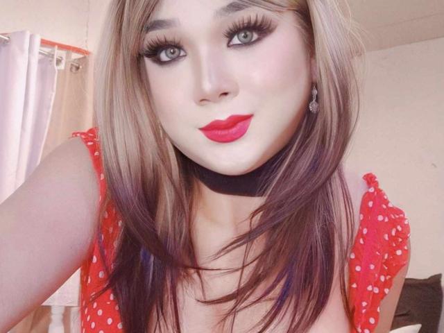 MissBigCockLucy with long brown hair, red lipstick, and thick eyelashes, wearing a red polka dot top, looking into the camera with a slight pout. Her tits are partially visible.