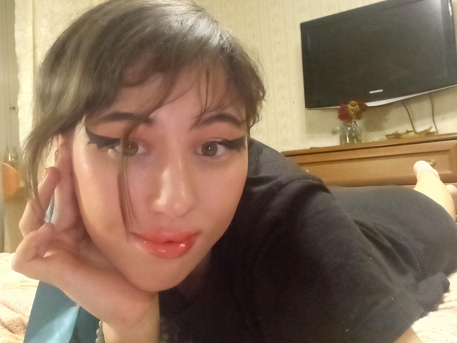 RowenaVonnie, laying on her stomach, black shirt, shiny red lips, long eyelashes, brown eyes, black hair, hand on cheek, flats in background, flat-screen TV on wooden dresser.