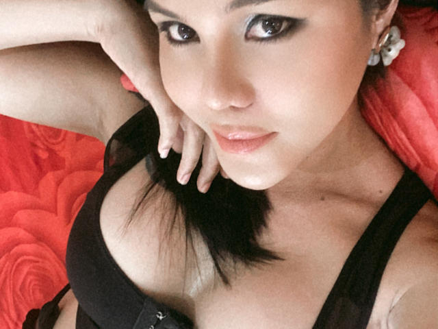 MajesticTrannyCock con delineador negro, labios brillantes y un sujetador negro, mostrando el escote y con una mano en el cuello, tiene cabello negro, aretes y una sonrisa seductora..