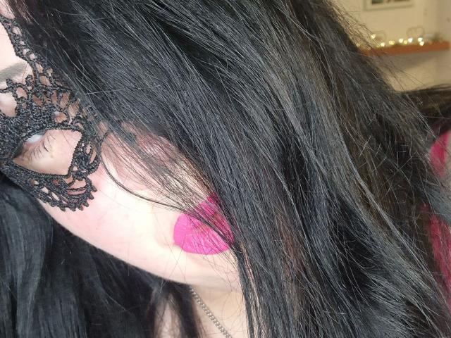 Blackbeauty43-hot's face with black lace eye mask, long black hair covering eyes, pink lipstick, and a hint of a silver necklace. Boobs partially visible, red clothing in background.