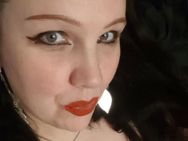 Blackbeauty43-hot's face with pale skin, red lipstick, black wavy hair, and black eyeliner, looking slightly up with blue eyes. She has a chain necklace and a subtle smile.