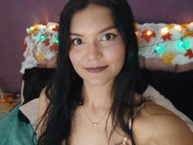 AriaLeenn has long black hair, dark brown eyes, and is smiling with brown lipstick. She's wearing a necklace and a floral top, showing her tits slightly.