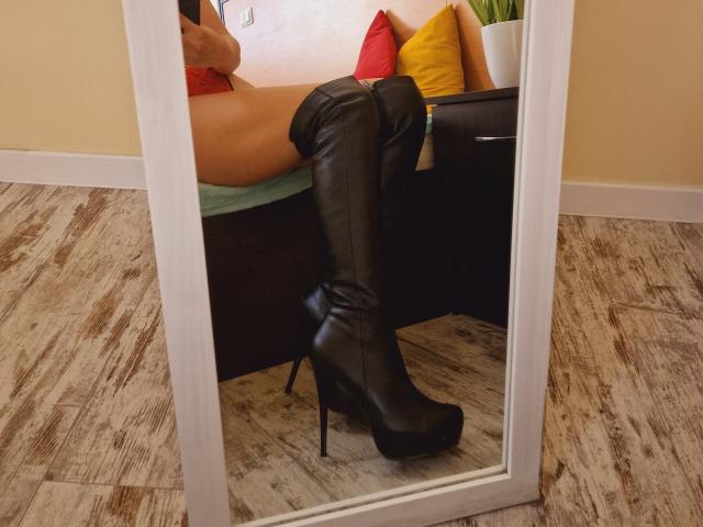 Les bottes en cuir noir montantes jusqu'à la cuisse de Jumiera, avec des talons hauts, se reflètent dans un miroir. Ses fesses sont partiellement visibles, et elle porte de la lingerie rouge. Le sol est en bois, et le miroir est blanc..