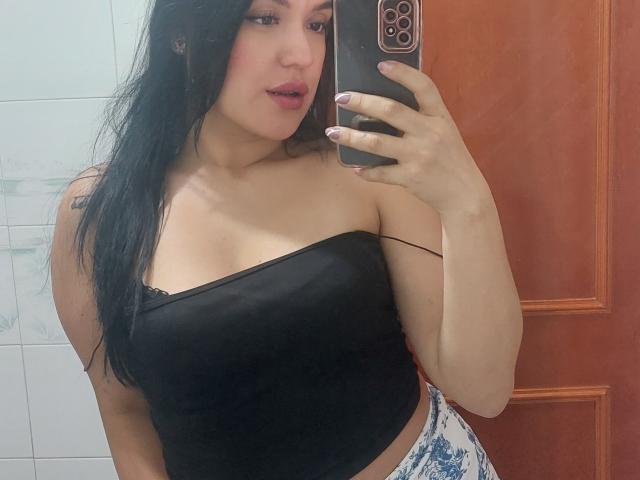LauraRichis with long black hair, pink lipstick, black strapless top showing a little black lace, holding a phone, taking a mirror selfie, showing her tits and tattooed arm.