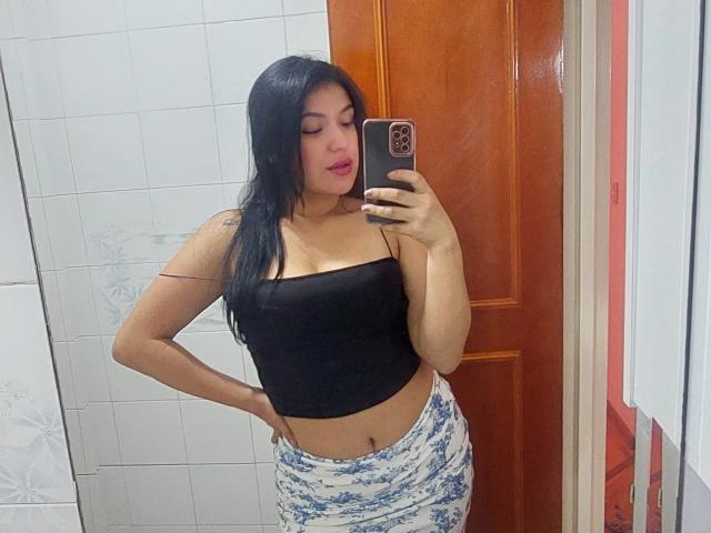 LauraRichis with long black hair, wearing a black crop top and blue floral skirt, holding a phone, taking a mirror selfie, showing her tits and a tattoo on her arm.