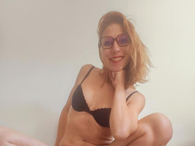 ApoloniaRed with red hair, glasses, and black bra, smiling, touching chin, sitting with legs bent, showing her small tits and ass, on a white background.