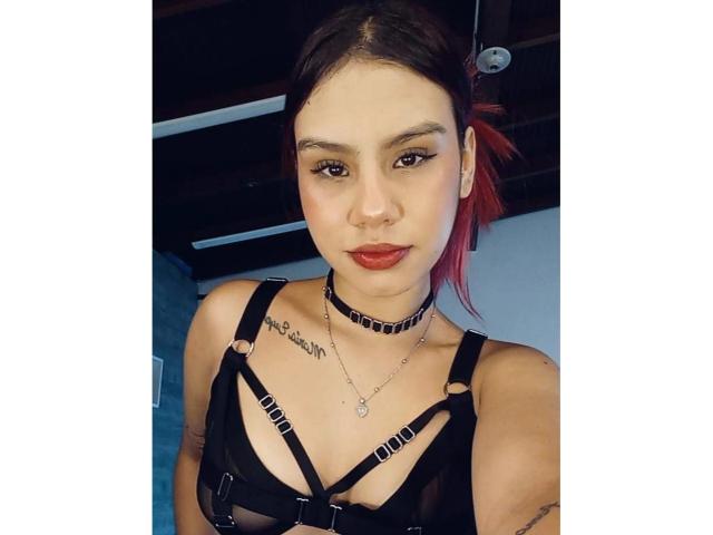 SussyGray with red-dyed hair, black strappy top, choker, tattoos, red lipstick, and eyeliner, showing tits, looking at camera. Sassy, confident, and stylish.