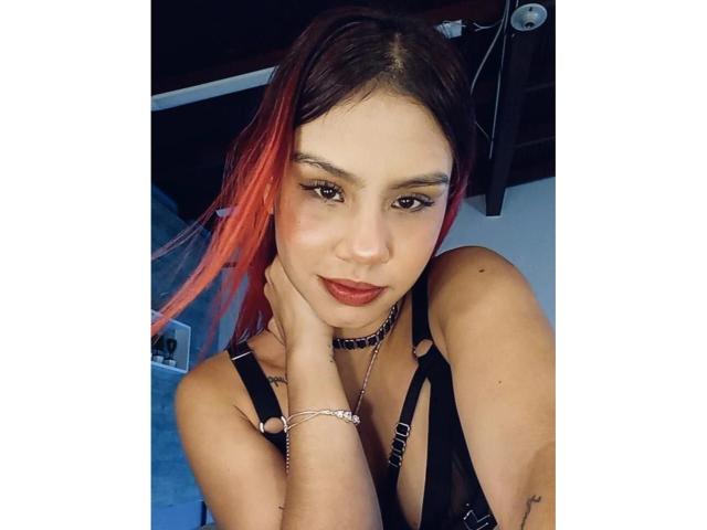 SussyGray has red-dyed hair, wearing black harness lingerie, choker, and bracelet, with dark makeup and red lipstick, resting her chin on her hand, looking at the camera.