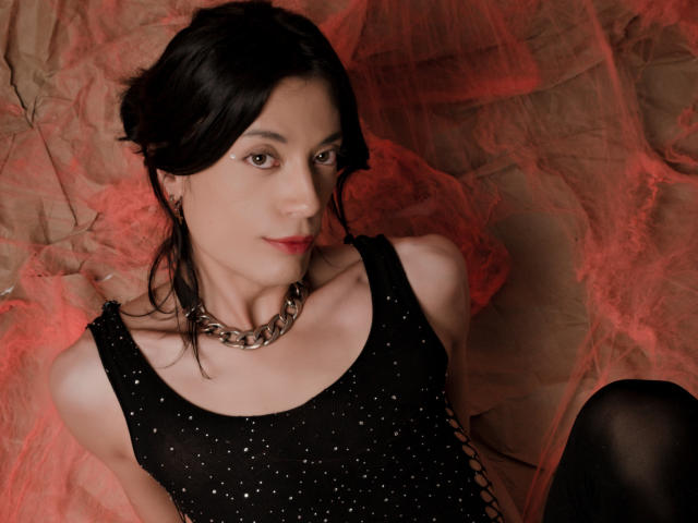 MilarHole with black hair, wearing a sparkly black dress, chunky chain necklace, and black stockings, sitting on red and beige fabric. Boobs visible, looking at camera.