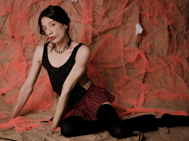 MilarHole with pale skin, black hair, wearing a black sparkly top, red plaid skirt, black stockings, and a chunky necklace, sitting on brown fabric with red netting.