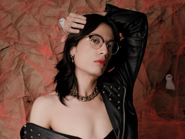 MilarHole with glasses, black leather jacket, and chain necklace, raising arm, exposing bare shoulders, red lipstick, and earrings, against crumpled brown paper with red webbing.