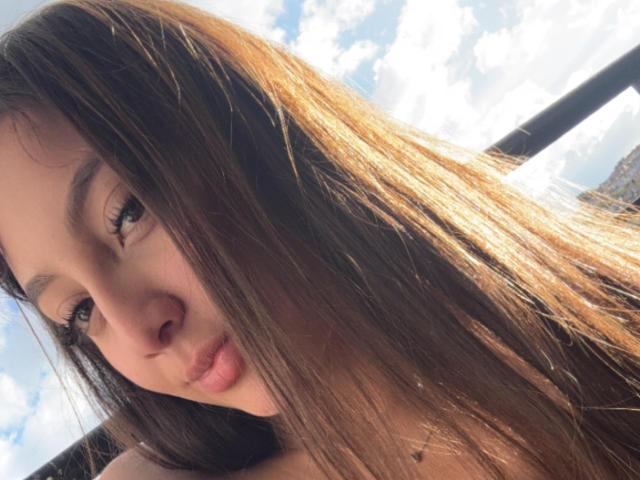 Le visage de DianaFernandezz, avec ses longs cheveux bruns, ses lèvres pulpeuses et ses yeux marron, la lumière du soleil dans ses cheveux, un ciel bleu avec des nuages en arrière-plan. Ses fesses ne sont pas visibles..
