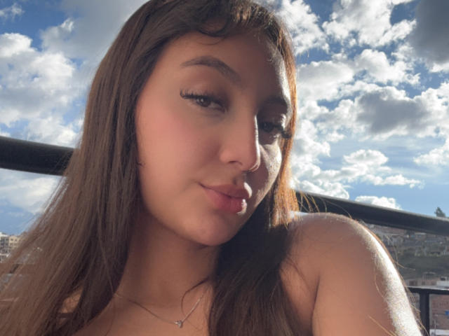 DianaFernandezz has long brown hair, dark eyeliner, and a subtle smile with sunlight on her face, showing her bare shoulders. Her tits are slightly visible, and she's outside.