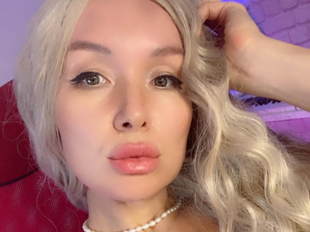 AvaGreenx heeft blond, golvend haar, volle, roze lippen en een parelketting, met een hand in haar haar, waardoor haar schattige, sexy gezicht met subtiele make-up goed te zien is..