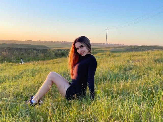 AliceWeiss con lunghi capelli castani, una maglia nera a maniche lunghe e pantaloncini neri, seduta sull'erba verde, sorridente, con le gambe incrociate, parte dei glutei visibile, tramonto, cielo sereno, linee elettriche sullo sfondo..