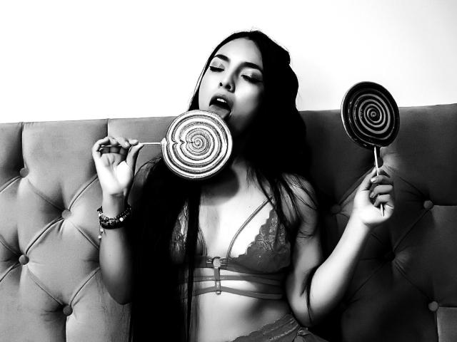 SakuraHarper, avec de longs cheveux noirs, un soutien-gorge en dentelle, tenant deux sucettes en spirale, une dans la bouche et une à la main, assise sur un canapé à capitonnage, avec une expression séduisante, photo en noir et blanc..