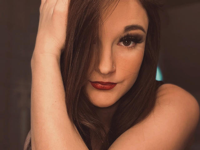 Scarlet-Young-hot has brown hair covering one eye, red lipstick, and a small nose piercing. Her arms are raised, hands in her hair. She has long eyelashes and a subtle smile.