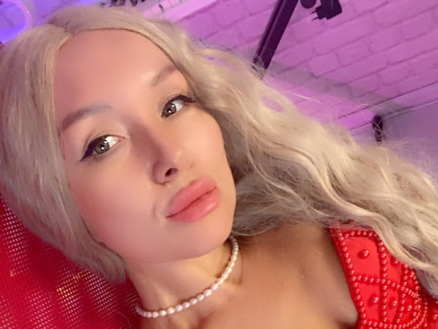 AvaGreenx with long, wavy blonde hair, pouty pink lips, and a pearl necklace, wearing a red dress with bead details, showing her boobs in a selfie.