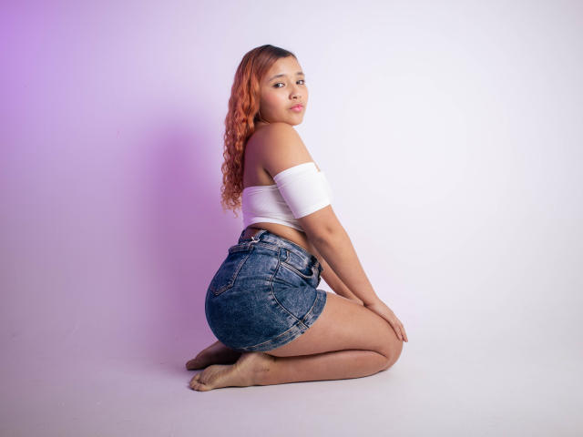 KarolAvalos, con i capelli ricci arancioni, inginocchiata, indossa una maglietta bianca a spalle scoperte e pantaloncini di jeans, mostrando il sedere, guarda indietro, a piedi nudi, su uno sfondo bianco con luce viola..