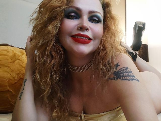 AnnaRichie with curly red hair, blue eyeshadow, red lipstick, nose ring, pearl necklace, tattoo on right shoulder, lying on bed, yellow pillow, black high heel in background.