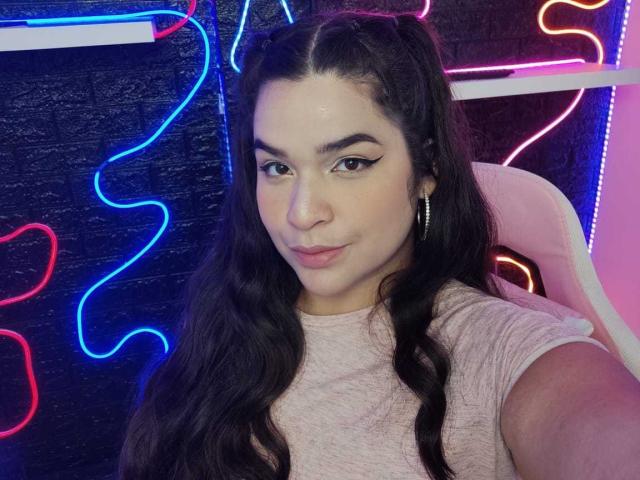 AntonelaPark with long black wavy hair, light pink shirt, hoop earrings, and subtle eyeliner, smiling slightly, neon blue and pink lights in background.