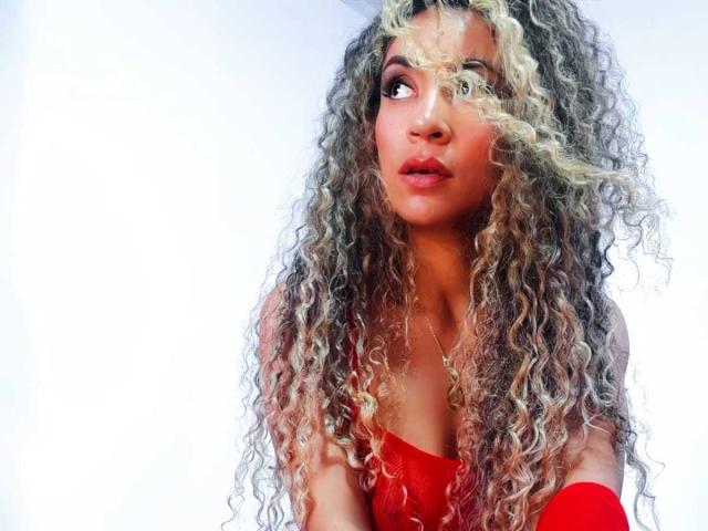 KylieJen with long, curly blonde and brown hair, wearing a red top, looking to the side with a slightly pouty expression, red lipstick, and a light brown skin tone.