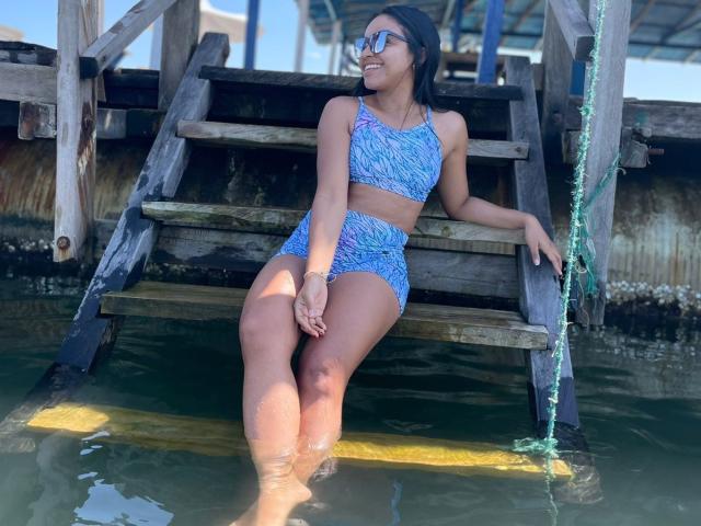 GiuLatina sitting on wooden stairs in water, wearing blue patterned bikini top and bottoms, sunglasses, and a wristwatch, legs in water, smiling, dark hair, tan skin.
