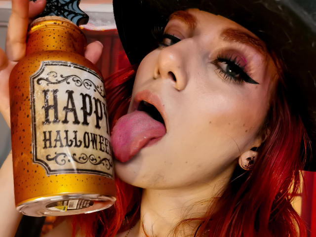 ZoeeMar with red hair, black eyeliner, and pink lipstick, tongue out, licking a pumpkin beer can, wearing a black witch hat, with a slightly open mouth, showing her tongue.