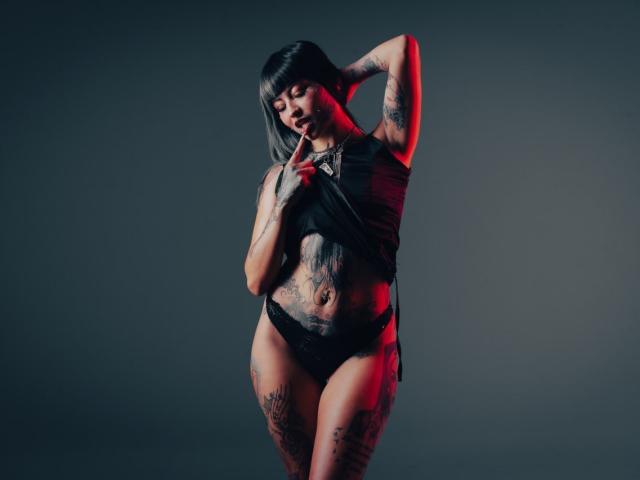AnaisMoon69 with tattoos, black top, and black panties, lifting her shirt, showing her tits and belly, right hand behind head, red and blue lighting, dark background.