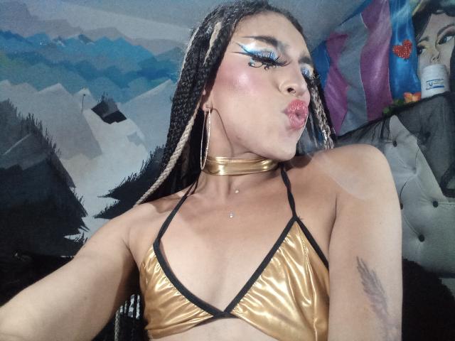 Crystalinn with gold metallic bikini top, black trim, choker, and long braided hair, pouting lips, bold blue eyeliner, and a tattoo on her right arm.