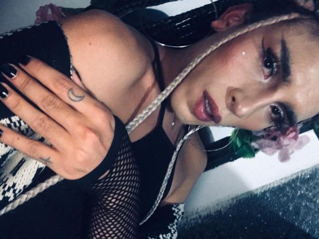 Crystalinn with braided hair, black fishnet arm sleeves, and pink flower, showing a crescent moon tattoo on her hand, looking at the camera with glossy pink lips.
